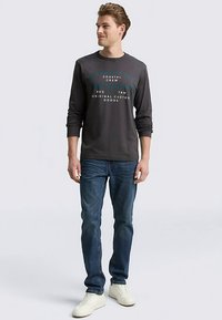 Grijs T-shirt met lange mouwen en een ronde hals met tekst, gecombineerd met een blauwe spijkerbroek en witte sneakers. Model staat voor een neutrale achtergrond.