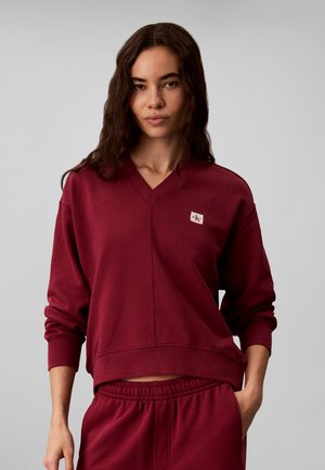Jonge vrouw met lang donker haar draagt bijpassende donkerrode trui en broek, staand met één hand in de zak tegen een effen achtergrond.