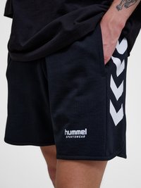 Person trägt schwarze Hummel-Sportshorts mit weißen Chevronstreifen und Logo, Hand ruht in der Tasche, sichtbares Tattoo am Arm.