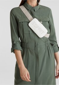 Bolso cruzado blanco con el logo "ESPRIT" en relieve, combinado con un vestido verde de botones que presenta una cintura entallada y mangas enrolladas.