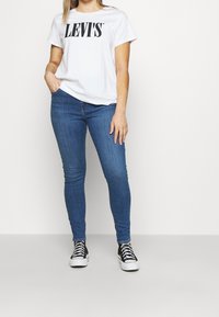 Bílá tričko s krátkým rukávem s logem "LEVI'S", v kombinaci s modrými skinny džínami a černými vysokými teniskami, stojící na neutrálním pozadí.