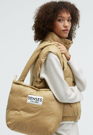 Beige puffer vest with high collar, worn over a light gray sweatshirt. The model carries a matching beige puffer tote bag with a logo.