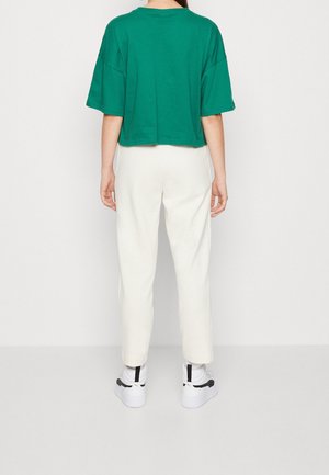 Person trägt ein lockeres grünes Crop-T-Shirt, weiße gerade Hose und weiße Sneakers mit schwarzen Akzenten, steht mit dem Rücken zur Kamera.
