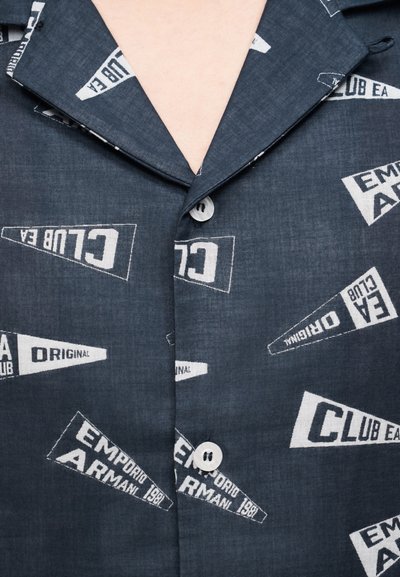 Σκούρο μπλε πουκάμισο με κουμπιά, διακοσμημένο με λευκές εκτυπωμένες σημαίες που φέρουν κείμενα όπως "CLUB EA" και "EMPORIO ARMANI 1981."
