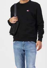 Svart sweatshirt med rund halsringning, prydd med en liten färgglad logotyp på bröstet, ihop med ljusblå jeans och en svart axelremsväska.