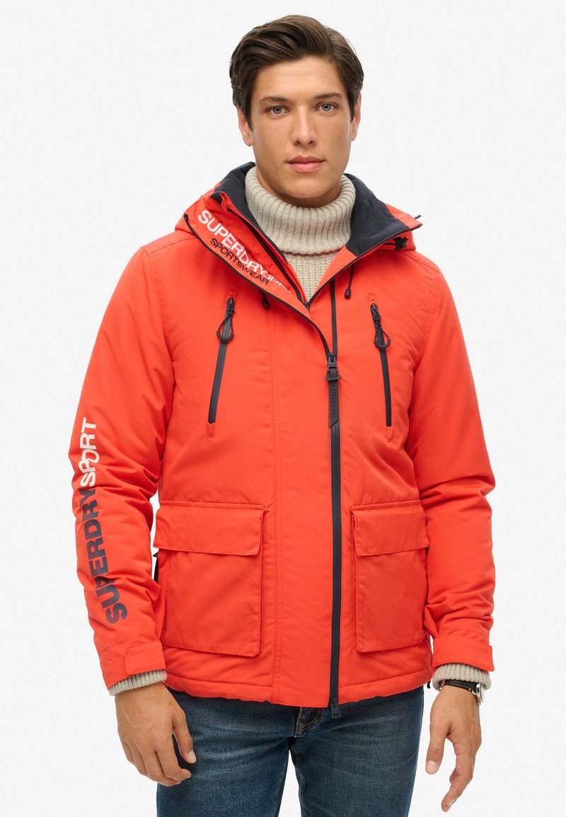 Chaqueta naranja brillante con capucha, cierre frontal y dos bolsillos en el pecho. Presenta acentos negros y un logo en la manga. Lleva puesta sobre un cuello de tortuga beige.