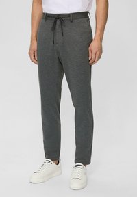 Graue Sweatpants mit einem Kordelzugbund, schmalem Schnitt und glatter Textur. Tragen mit weißen Sneakers vor einem neutralen Hintergrund.