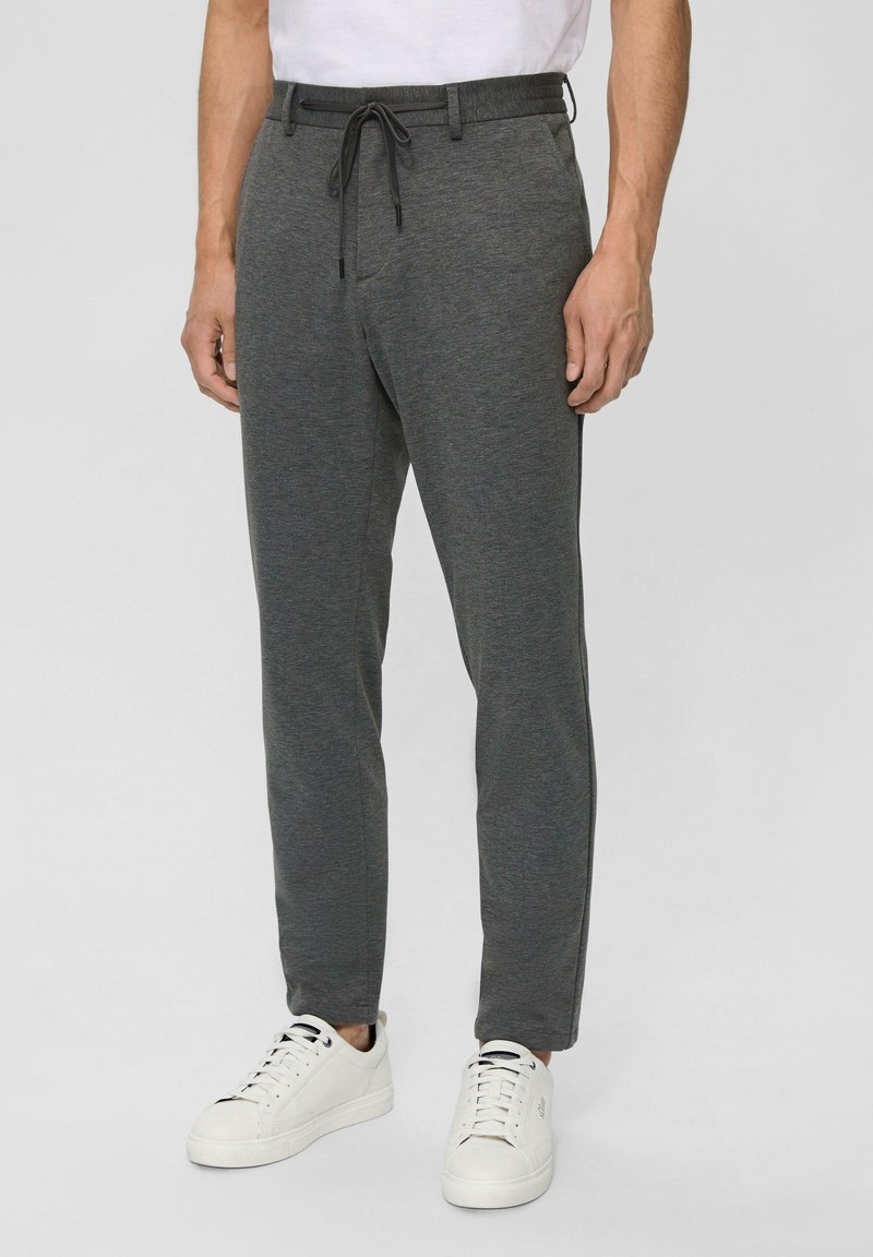 Graue Sweatpants mit einem Kordelzugbund, schmalem Schnitt und glatter Textur. Tragen mit weißen Sneakers vor einem neutralen Hintergrund.