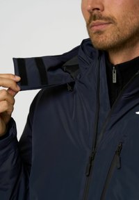 Mann, der den Kragen einer marineblauen Jacke mit Klettverschlüssen anpasst, während die Jackenreißverschlüsse und die schwarze Innenschicht gezeigt werden.
