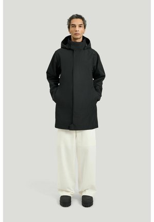 Person med krøllet hår iført en sort hættetrøje, hvide bukser med vide ben og sorte chunky slip-on sko, der står mod en ensfarvet baggrund.