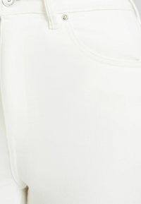 Jeans de mezclilla blanca que presentan una vista en primer plano de la tela suave, costuras bordadas y acentos de hardware metálico en la cinturilla.