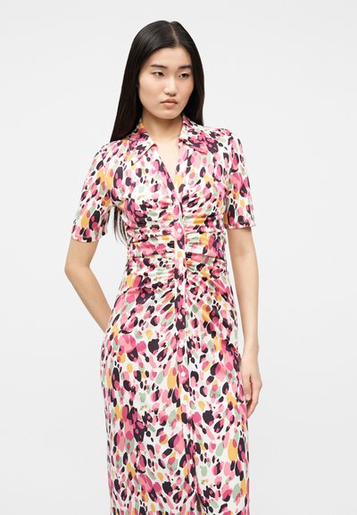 Femeie cu păr lung și închis la culoare, purtând o rochie midi colorată cu pete, cu nasturi, pliseuri, mâneci scurte și guler.