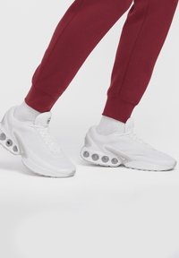 Person wearing white athletic sneakers with visible cushioning soles and maroon joggers, standing on a white background.