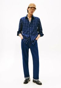Joven con gorra beige, camisa de cuadros azul de botones, jeans azules con puños y mocasines negros, de pie con las manos en los bolsillos.