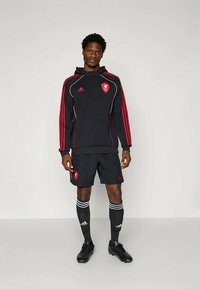 Felpa sportiva nera con strisce rosse e logo, abbinata a pantaloni corti neri, calze lunghe fino al ginocchio con strisce bianche e scarpe da calcio nere.