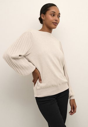 Pull en maille beige avec des détails côtelés sur les manches bouffantes, col rond et texture douce. Associé à un pantalon foncé.