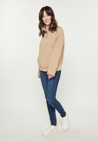 Beige steppelt pulóver magas gallérral és elülső zsebbel, sötétkék farmerrel és fehér sportcipővel párosítva, mely egy laza öltözetet alkot.