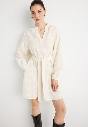 Φόρεμα εκρού με υφασματοποιημένο λουλουδάτο μοτίβο, V-λαιμόκοψη, μακριά φουσκωτά μανίκια και ζώνη στη μέση. Συνδυασμένο με μαύρες μπότες μέχρι το γόνατο.