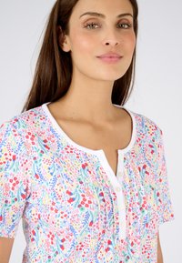 Korte mouwen, overhemd met knopen en bloemenpatroon in rood, blauw en groen op een witte achtergrond. Zachte stof met een ronde halslijn.