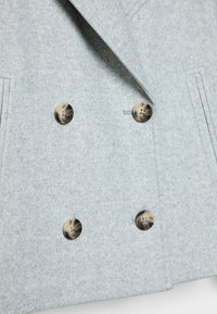 Manteau gris croisé en tissu feutré doux. Il présente des boutons bruns remarquables, des revers structurés et des poches latérales.