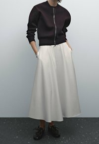 Schwarze Zip-Up-Sweatjacke mit Rundhalsausschnitt, kombiniert mit einem fließenden, knielangen weißen Rock. Das Modell trägt schwarze Schnürschuhe.