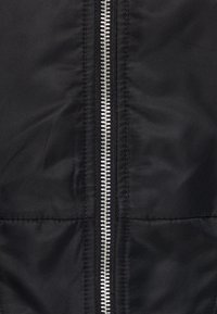 Svart jacka med en central silverfärgad dragkedja, quiltad textur och slät tygfinish. Inga synliga mönster eller extra detaljer.