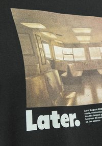 Sort t-shirt med et vintage-inspireret graphic af et toginteriør med sæder og vinduer, med ordet "Later." trykt i fede hvide bogstaver.