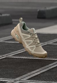 Sapatilha atlética bege com parte superior em malha texturizada, detalhes verdes e sola flexível. Design em zigzag notável e biqueira reforçada.