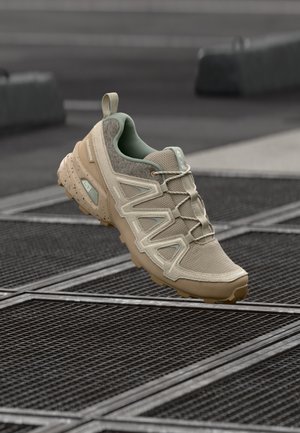 Sapatilha atlética bege com parte superior em malha texturizada, detalhes verdes e sola flexível. Design em zigzag notável e biqueira reforçada.