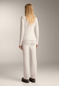 O persoană cu părul lung, ondulat și blond stă întoarsă, purtând un top cu mâneci lungi de culoare bej deschis, pantaloni asortați și pantofi cu platformă negri.