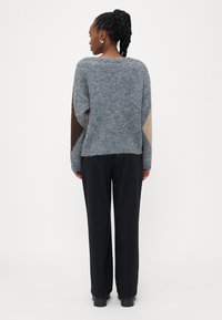 Pulover din tricot gros, gri, cu mâneci colorate în nuanțe de maro și bej, asortat cu pantaloni largi negri. Textură moale și croi lejer.