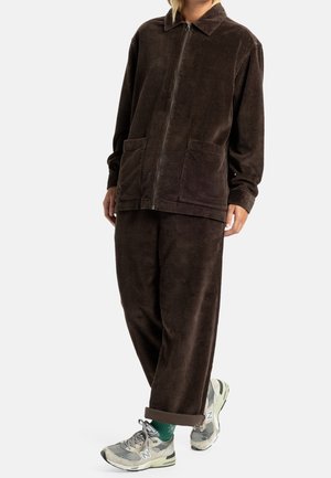 Person iført løs, mørkebrun ribstrikket lynlåstrøje og matchende bukser med grå og hvide sneakers og grønne sokker.