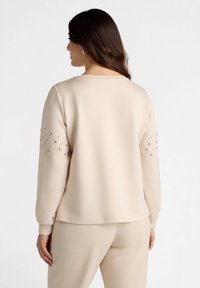 Beige sweatshirt with long sleeves, features decorative studs on the arms, round neckline, and textured fabric. Smooth finish and relaxed fit.