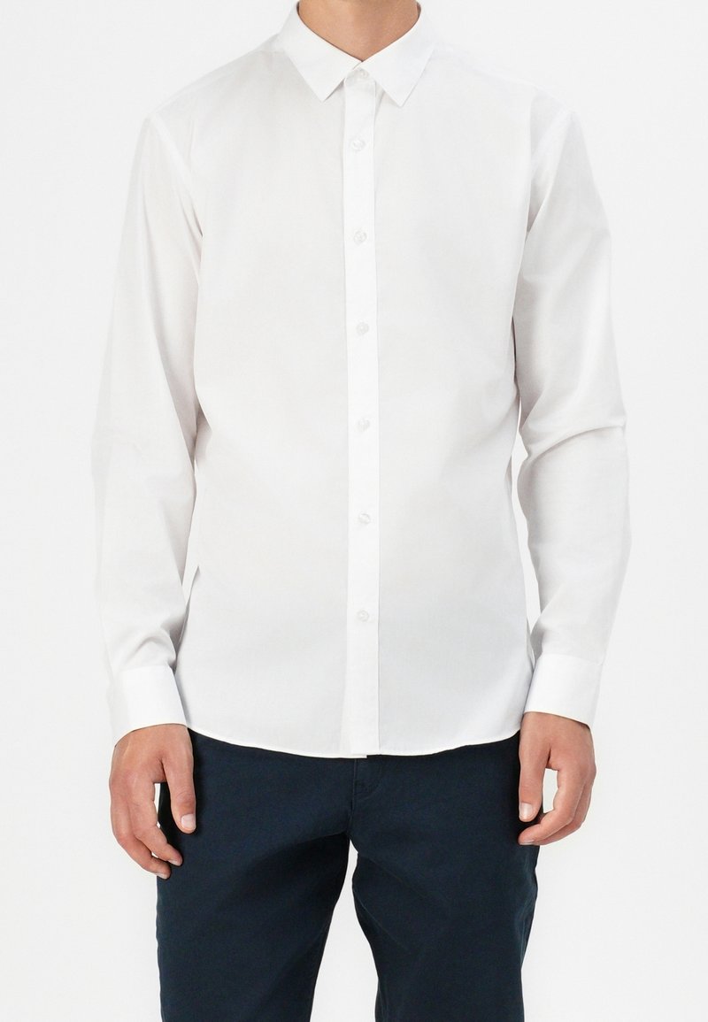Homme portant une chemise blanche à manches longues boutonnée et un pantalon bleu marine foncé, debout devant un fond blanc uni.
