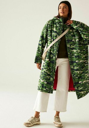 Kobieta w zielonym kamuflażowym długim płaszczu, białych spodniach 7/8, beżowych trampkach, ciemnym golfie, czarnej opasce na głowę i kremowej torebce na ramię.