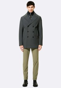 Cappotto peacoat doppiopetto grigio scuro con grandi bottoni, tasche laterali, indossato sopra una giacca nera con colletto alto e zip, abbinato a pantaloni verde oliva.