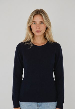 Vrouw met lang blond haar, een zwarte trui met lange mouwen en lichtblauwe spijkerbroek, staande tegen een effen lichte achtergrond.