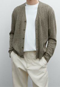 Brun, ribbad cardigan med knappar, bärs över en vit t-shirt. Tyget verkar strukturerat och kompletterar ljusa byxor.