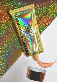 Gyllene tubförpackning med iriserande finish, märkt "24K Gold Lift & Firm Prism Cream." En persikofärgad kräm pressas ut på en vit yta.