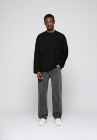 Black long-sleeve shirt with embroidered details on sleeves, paired with loose gray jeans and white sneakers on a plain background.
