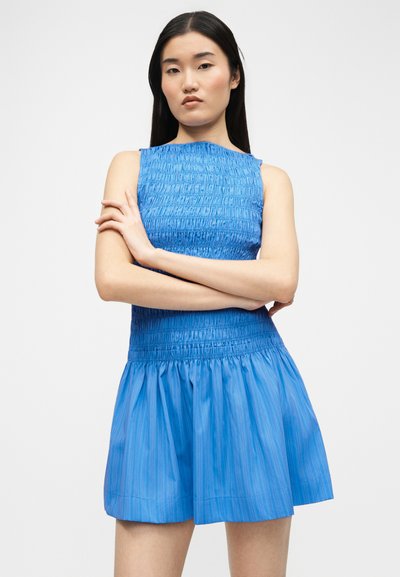 Νεαρή γυναίκα με μακριά σκουρόχρωμα μαλλιά, φορώντας μπλε αμάνικο φόρεμα με υφή, με σταυρωμένα χέρια, στέκεται μπροστά σε απλό λευκό φόντο.