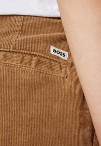 Gros plan sur un pantalon en velours côtelé marron avec une petite étiquette blanche portant la mention BOSS près d'une poche arrière, à côté du bras et de la chemise blanche d'une personne.