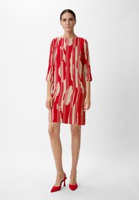 Rotes und beiges gemustertes Kleid mit Dreiviertelärmel, einem Bootsausschnitt und geradem Schnitt. Kombiniert mit roten High Heels.