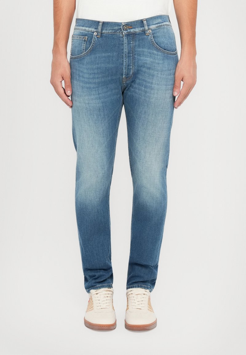 Ανοιχτό μπλε τζιν παντελόνι με ίσια γραμμή, που δ ιαθέτει πέντε τσέπες, κουμπί και ελαφριά φθορά στα πόδια. Φοράει μπεζ αθλητικά παπούτσια.