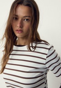 Ung kvinde med langt brunt hår iført en hvid skjorte med mørke, vandrette striber, der kigger let til siden mod en ensfarvet baggrund.