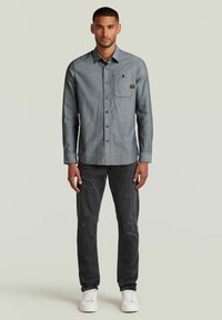 Chemise grise à boutons avec une poche, associée à un jean noir déchiré et des baskets blanches. Matériau en coton, manches longues, col classique.