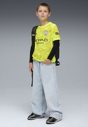 Maglietta da calcio gialla a fantasia con dettagli neri, jeans ampi azzurri chiari e sneakers nere. Il bambino porta uno zaino nero.