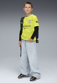 Maglietta da calcio gialla a fantasia con dettagli neri, jeans ampi azzurri chiari e sneakers nere. Il bambino porta uno zaino nero.