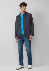 Grijze button-up jack over een blauw T-shirt, gecombineerd met blauwe jeans en witte sneakers. Het jack heeft twee voorkeurs.