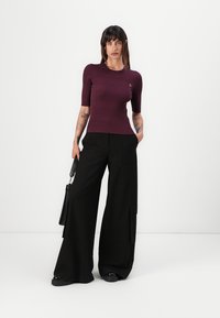 Bordeaux geribbelde top met korte mouwen, gecombineerd met zwarte pantalons met wijde pijpen en zijzakken; model houdt een zwarte handtas vast.